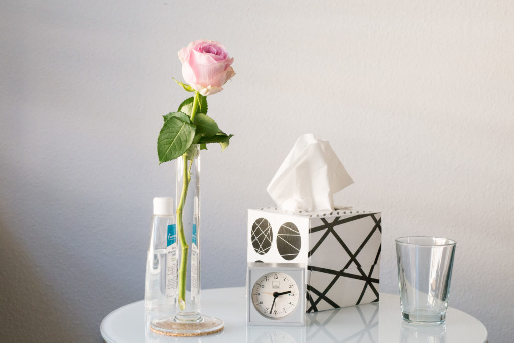 Ein Therapiebeistelltisch, auf dem eine Vase mit einer rosa Rose, eine kleine Uhr, eine Taschentuchbox und ein Glas Wasser steht.