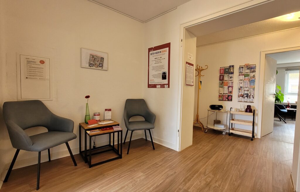 Ein kleiner Wartebereich mit zwei Stühlen. Poster und Informationsmaterial hängen an den Wänden.